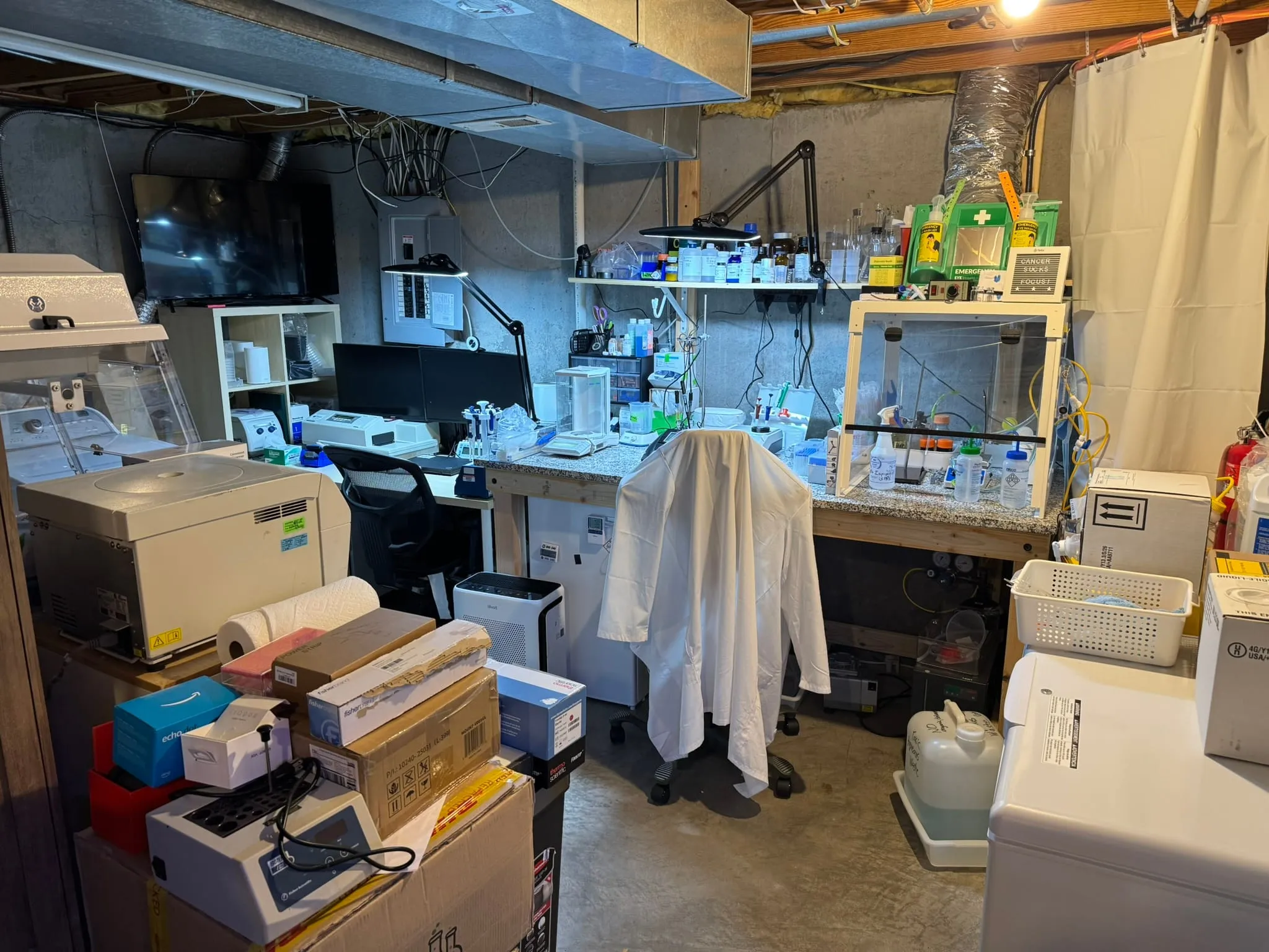 The basement laboratory where DuraRad began: granite-topped benches with an analytical balance, pipettes, a laminar-airflow enclosure, a reagent shelf with a 'Cancer sucks / Focus' sign, and a lab coat hung over the chair.