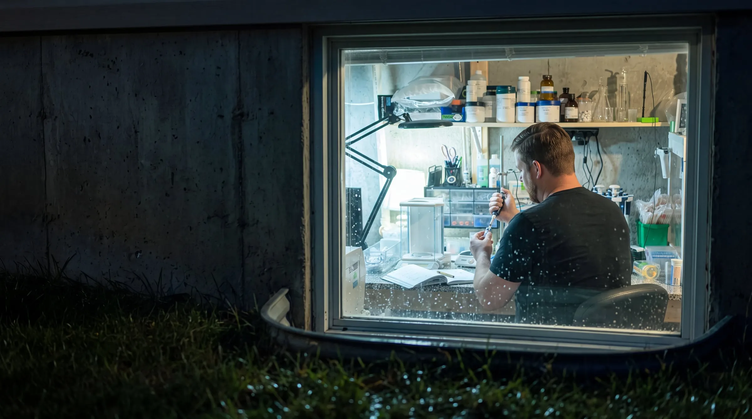 A lone scientist at work late at night in a cluttered basement laboratory, seen from outside through the lit, rain-flecked basement window — the origin of DuraRad.