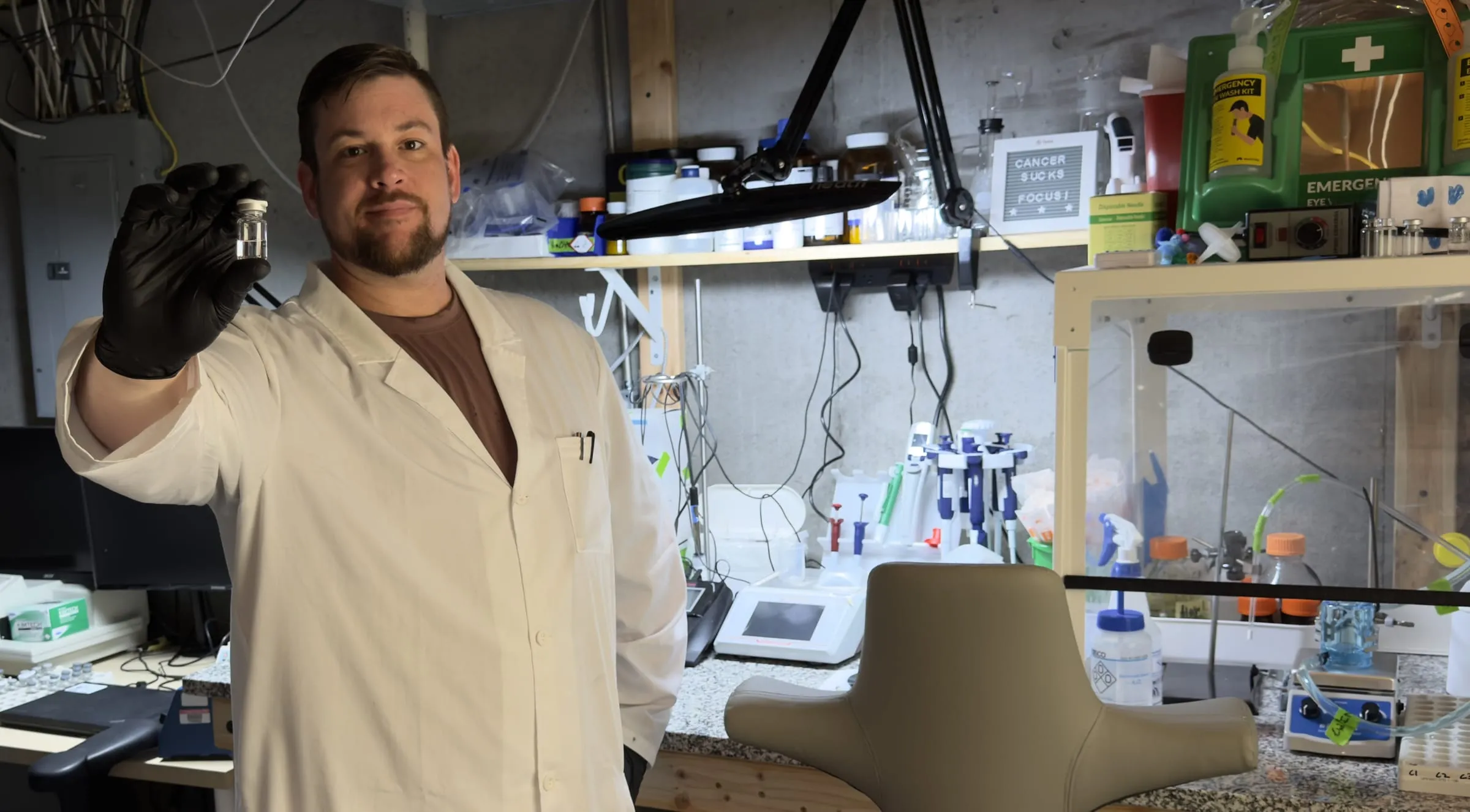 Chris Wiley in a white lab coat and black nitrile glove, holding a DuraRad-coated vial up to the camera at the basement-lab bench, with the reagent shelf and laminar-airflow enclosure behind him.