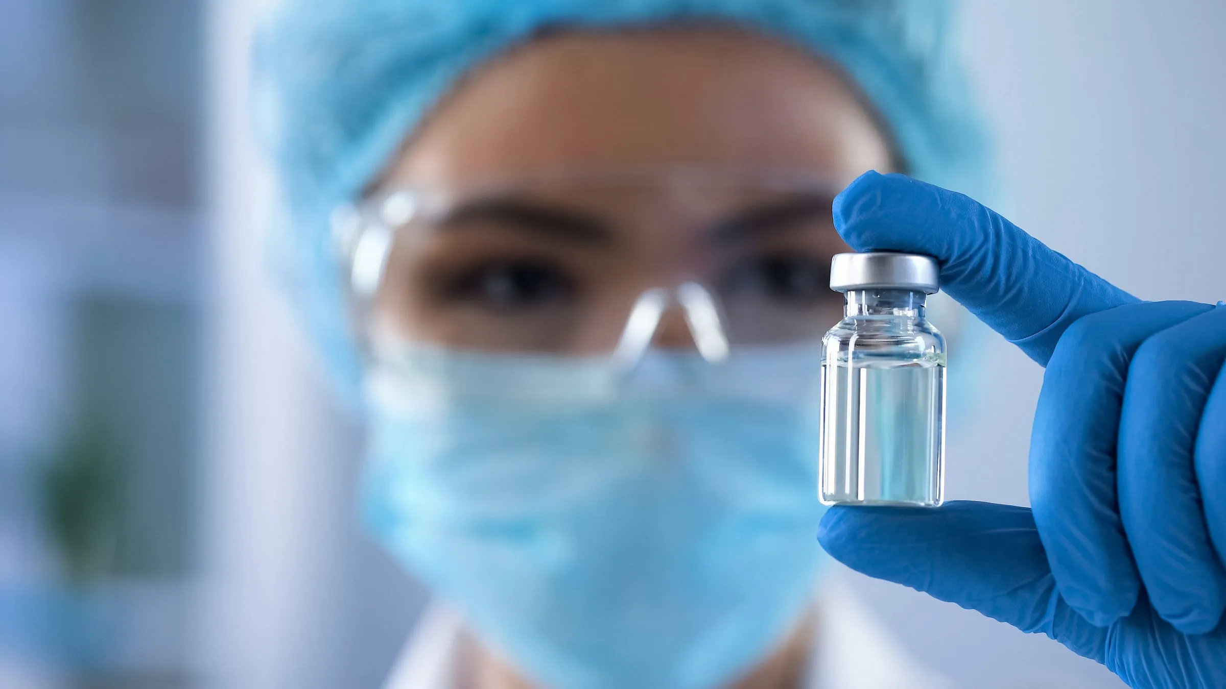 A gowned, gloved healthcare professional holding up a small clear glass vial to inspect it.