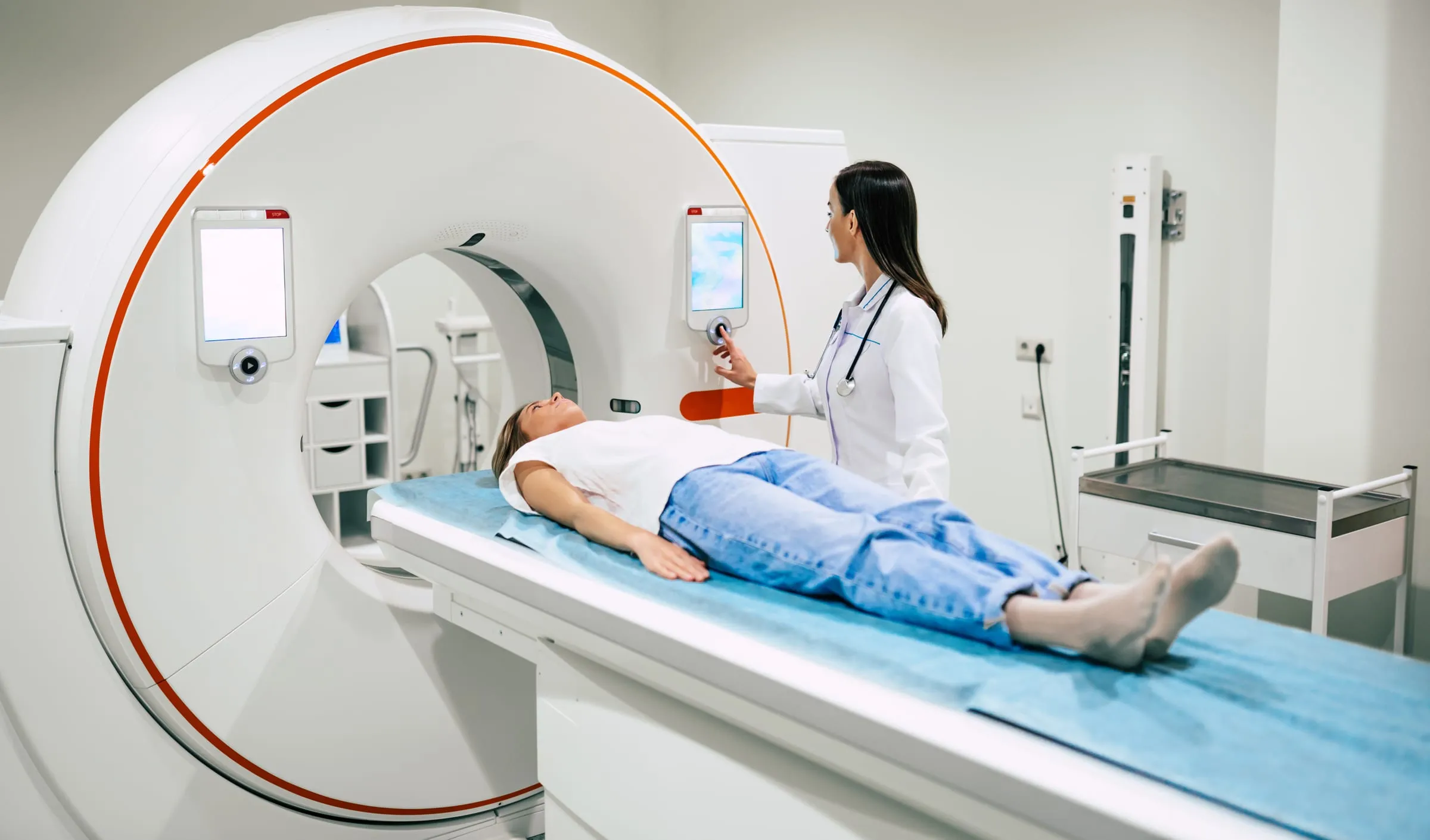 A patient lying on the table of a PET/CT scanner while a clinician in a white coat operates the machine.