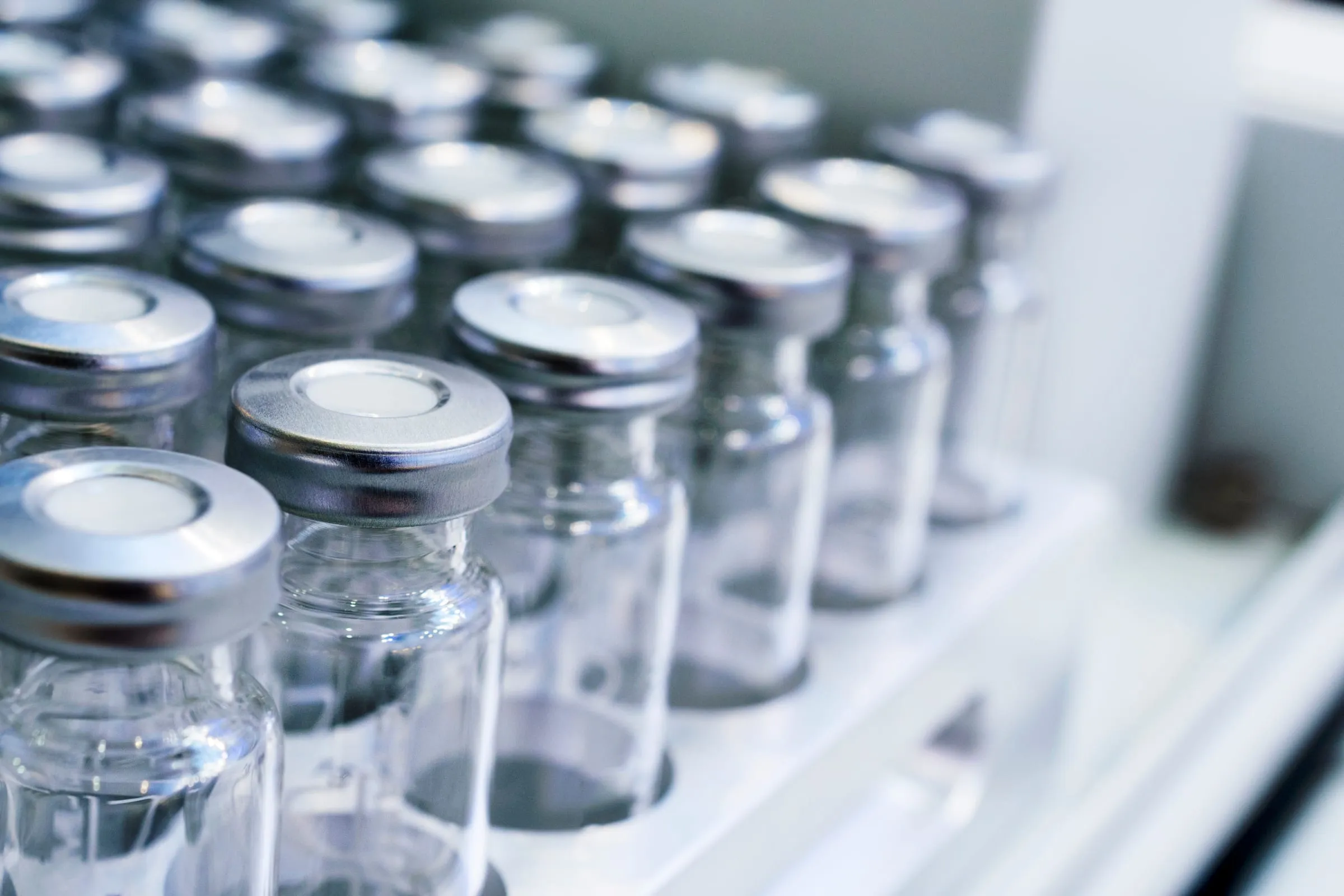 Rows of clean, empty clear glass serum vials with aluminum crimp seals in a tray, shallow depth of field, cool light.