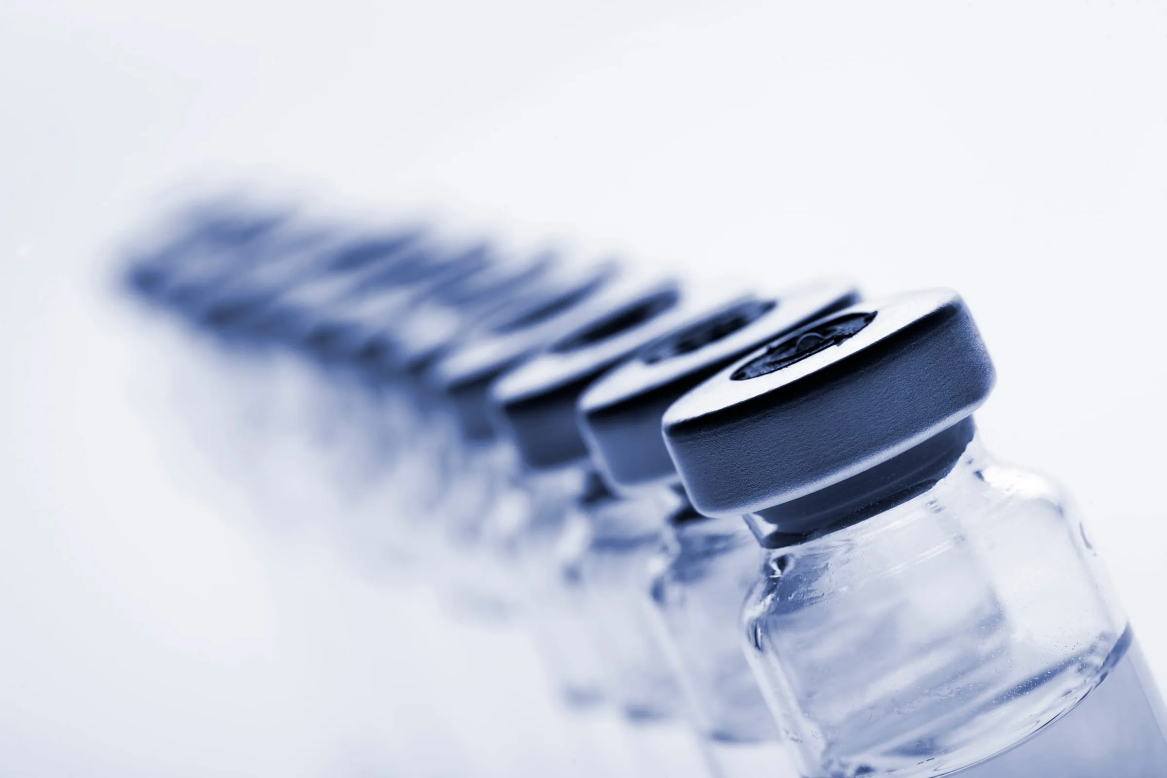 A single-file line of clear glass pharmaceutical vials with crimp seals, receding into soft focus on a bright, cool background.